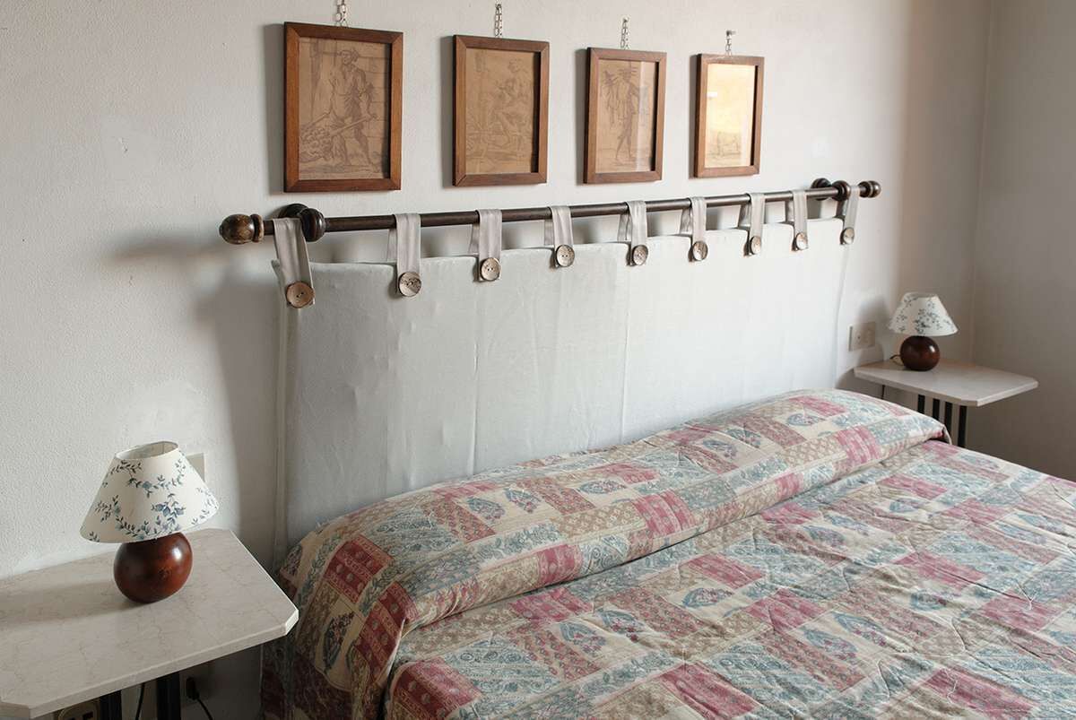 Une chambre à coucher avec un lit, une table de chevet avec une lampe, une tringle à rideaux fixée au mur et des œuvres d’art encadrées au-dessus du lit.