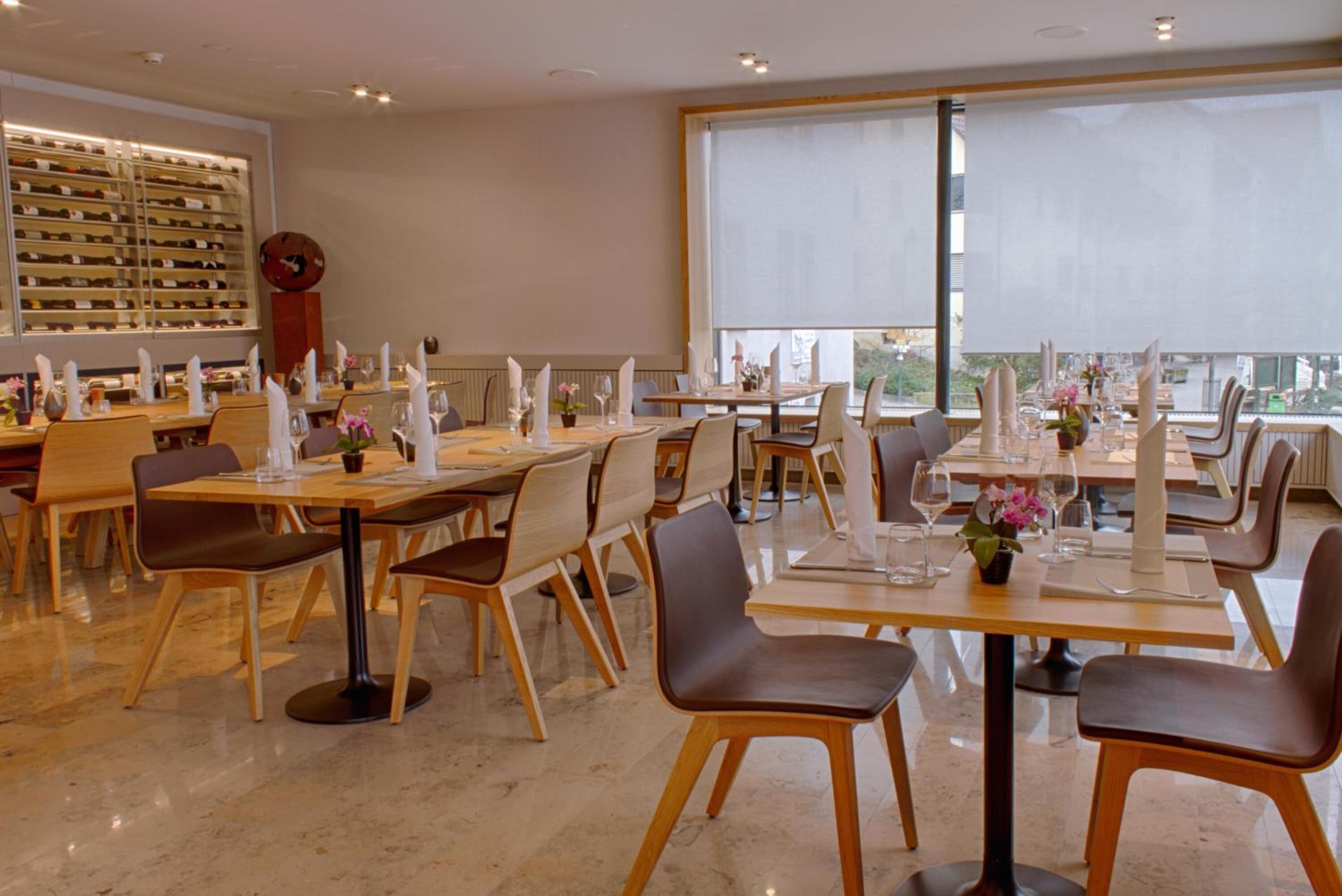 A modern dining area with multiple tables and chairs, large windows, and a wine rack in the background.