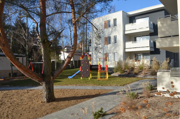 A playground with climbing and slide structures, surrounded by a building with multiple balconies and a tree. - Apartamenty na skarpie (Powidz)