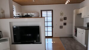 Un salon moderne doté d’un téléviseur à écran plat, de planchers en bois et d’un coin cuisine avec des meubles blancs.
