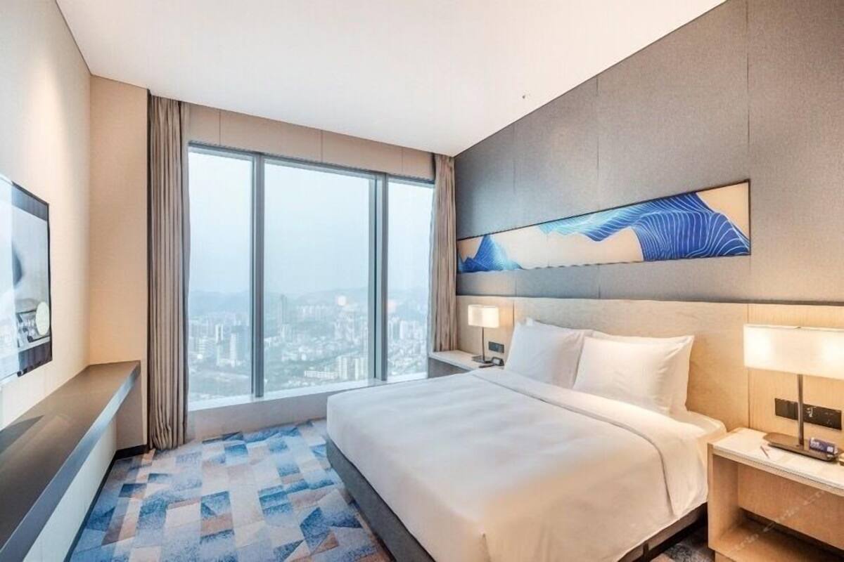 A modern hotel room with a large bed, a city view, and a painting on the wall.
