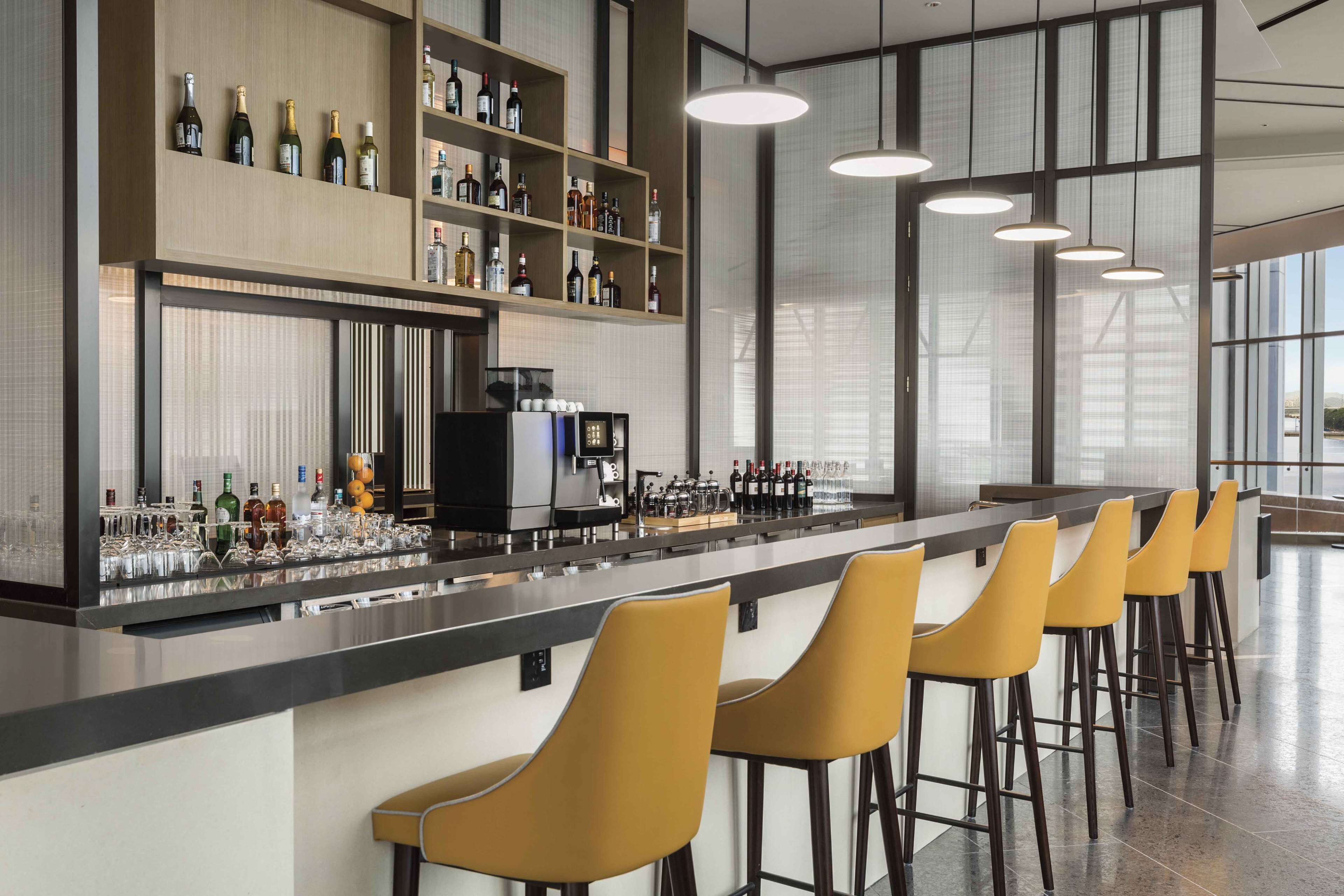 A modern bar area with a long counter, multiple shelves of bottles, and a coffee machine.