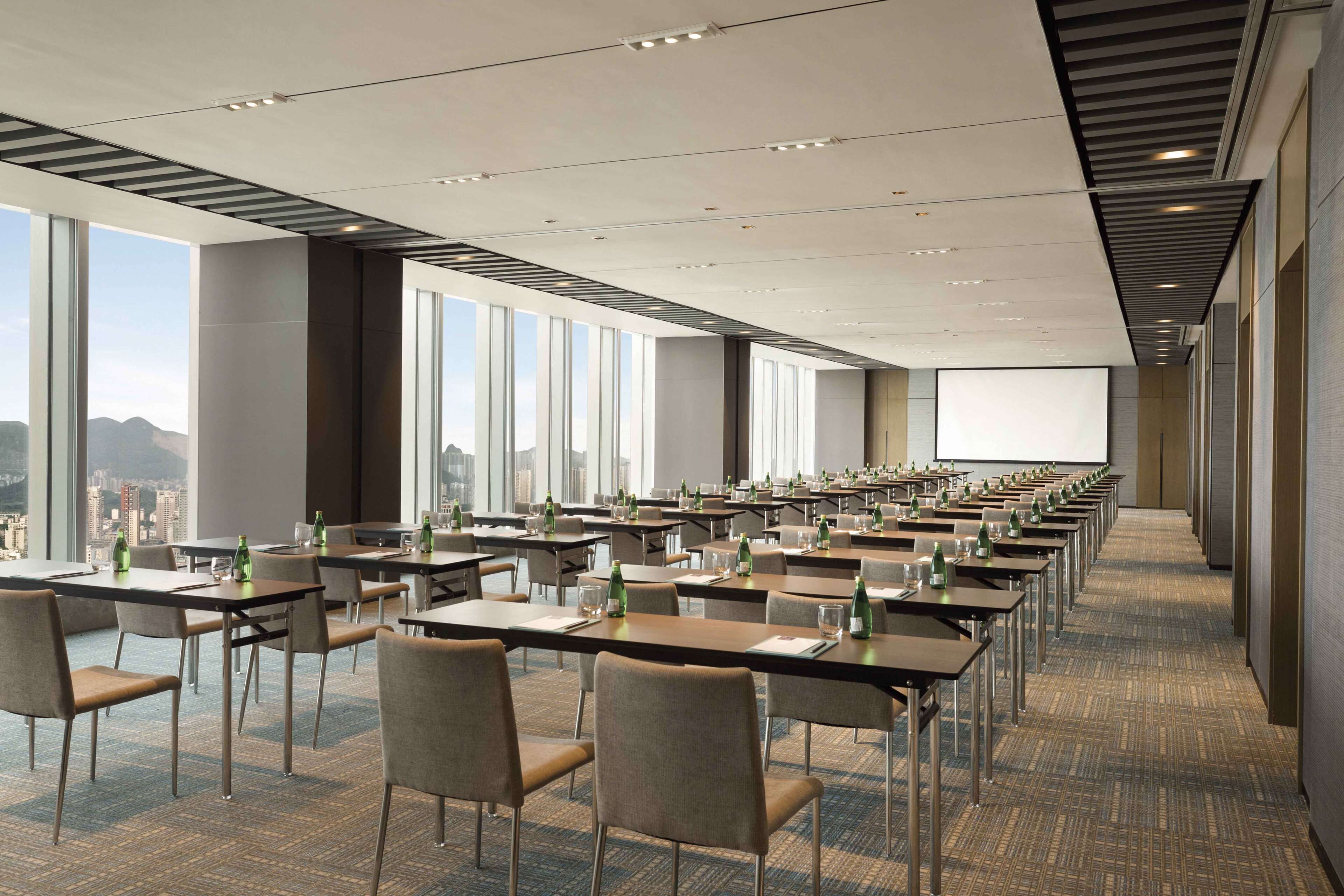 A conference room with rows of tables and chairs, large windows, and a projection screen.