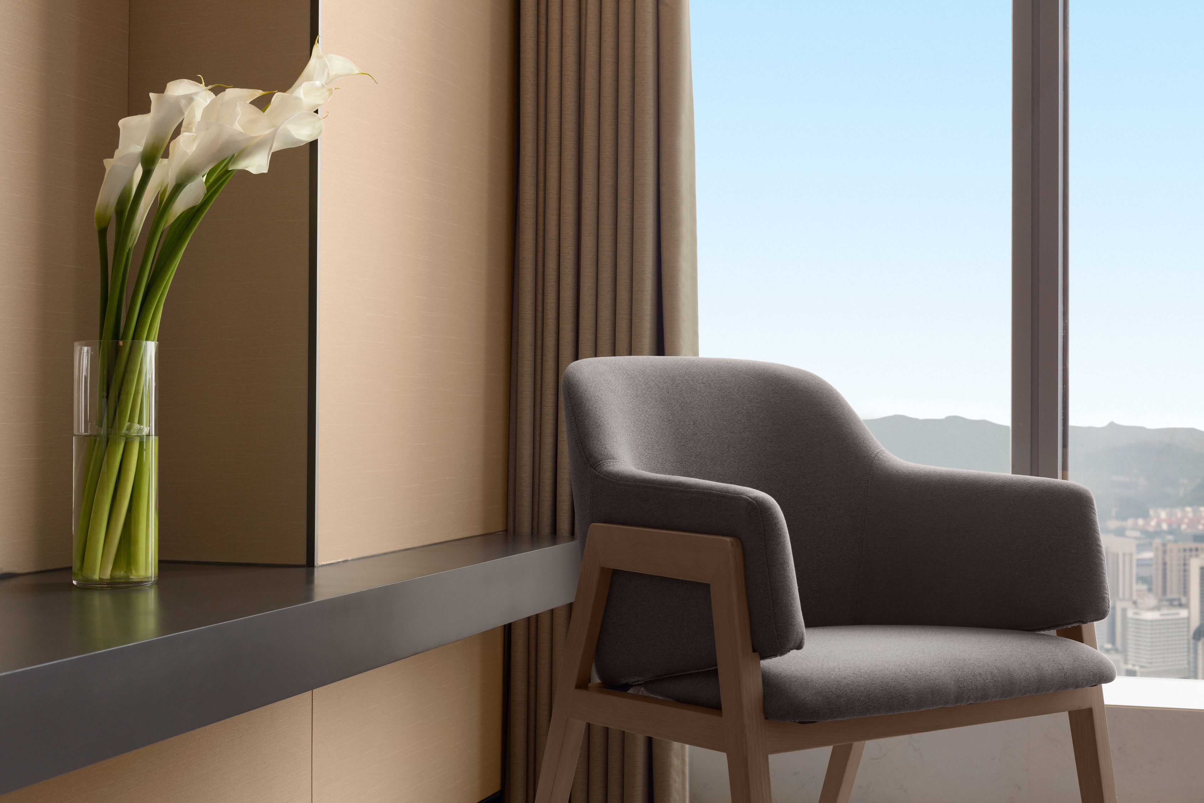 A modern room with a grey upholstered chair, a clear vase with white flowers, and a view of a cityscape through large windows.