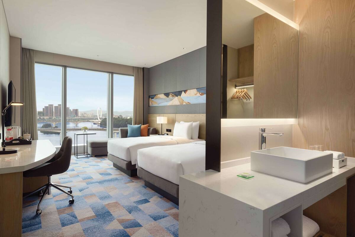 A hotel room with two beds, a desk, a large window with a city view, a bathroom with a large sink, and a painting on the wall.