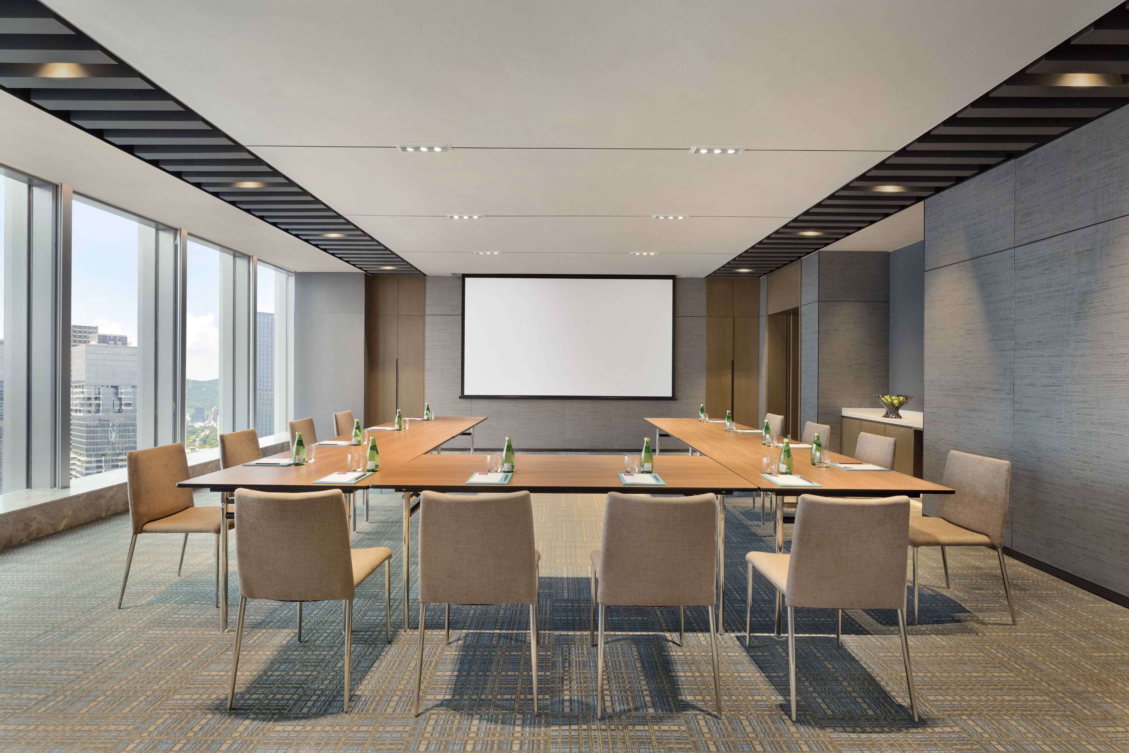 A conference room with a large table, chairs, a projector screen, and large windows offering a city view.