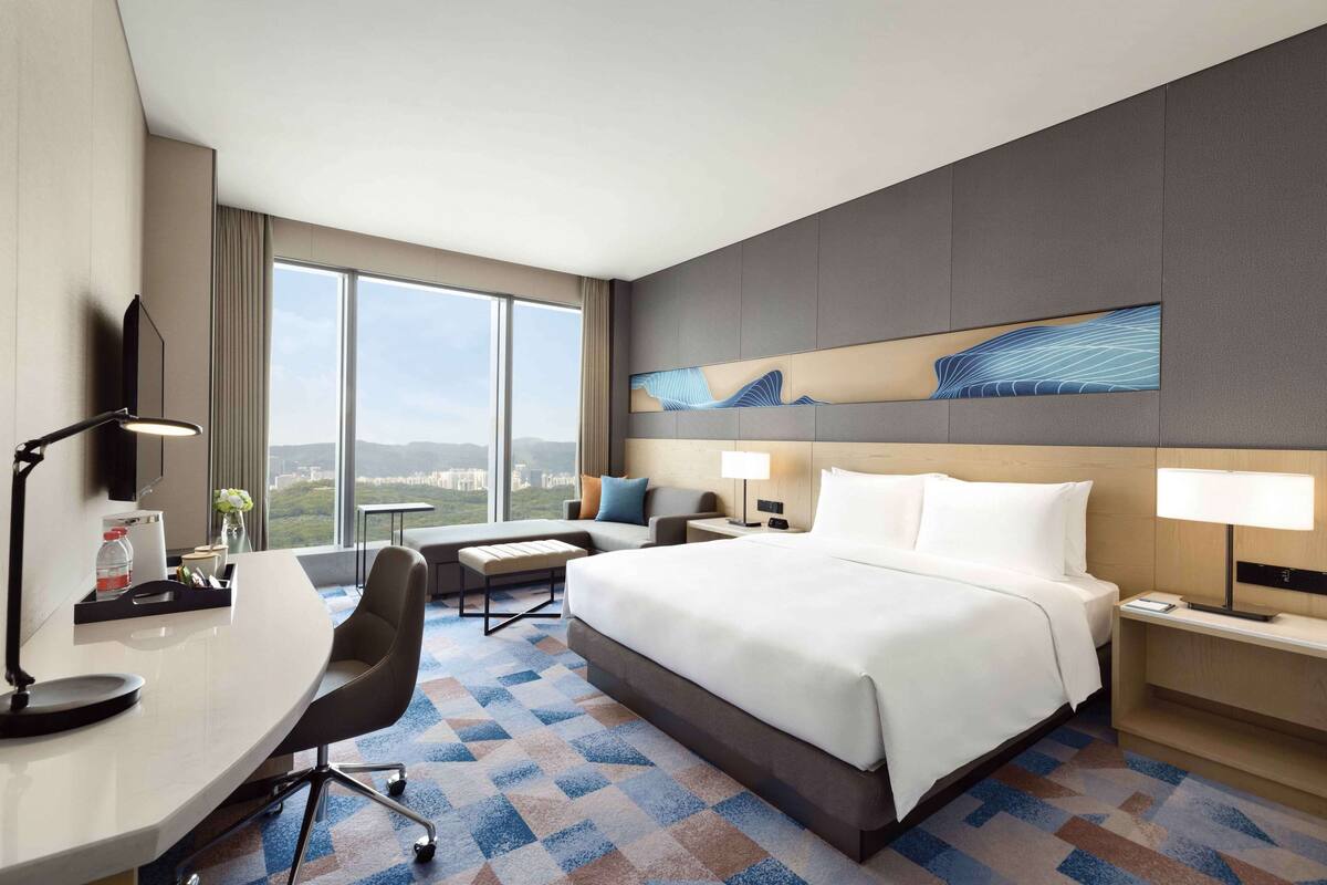 A hotel room with a large bed, a desk, a chair, a nightstand, and a wall-mounted TV. The room has a large window offering a view of the city.
