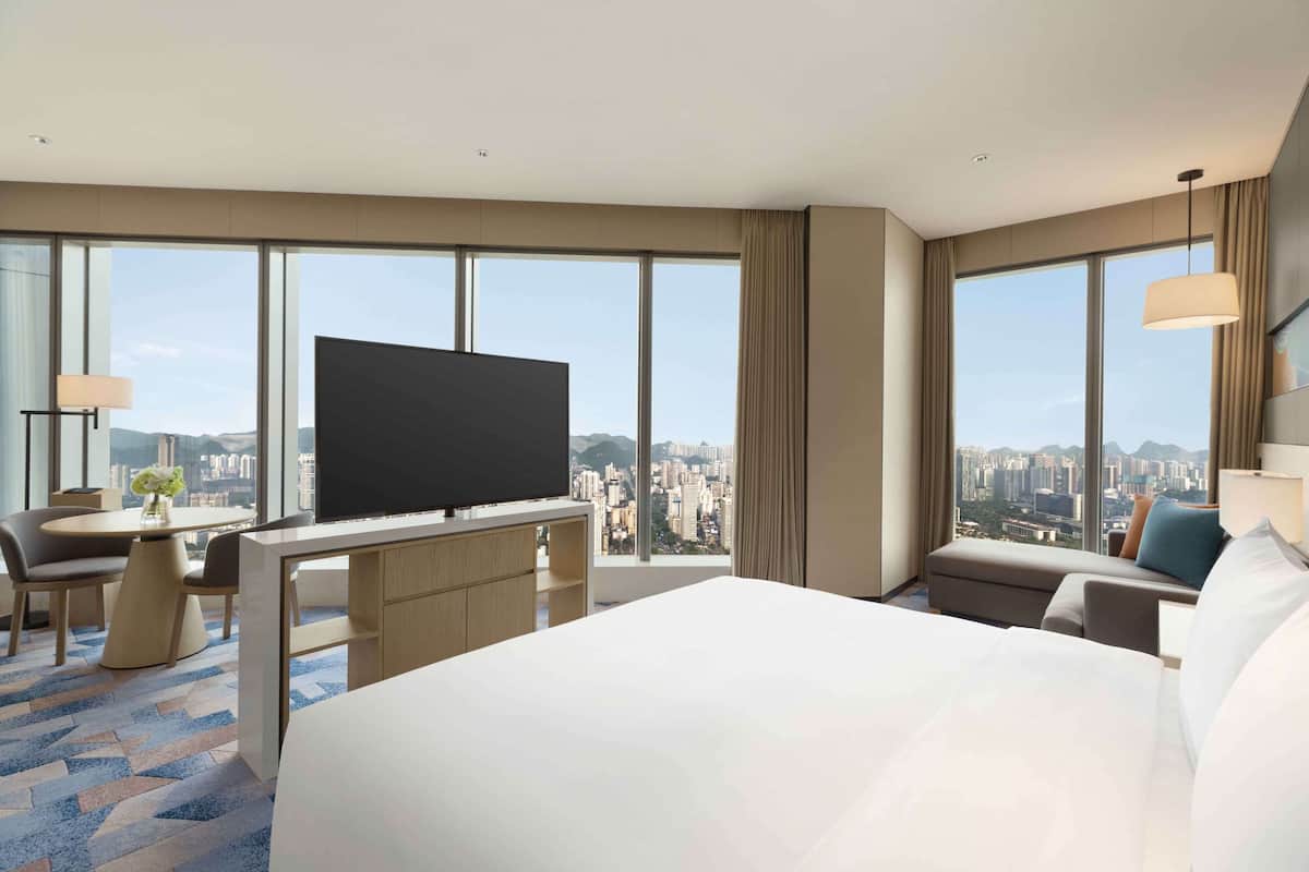 A hotel room with a large bed, a desk with a flat-screen TV, a seating area with a sofa and armchair, and a city view through large windows.