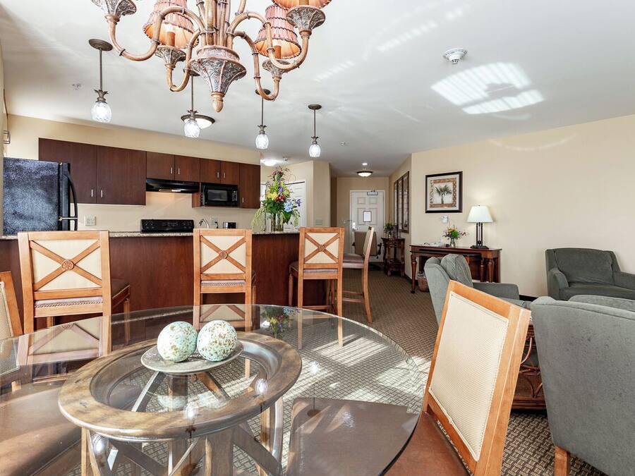 A spacious living area with a round glass table, wooden chairs, a green sofa, and a kitchen in the background.