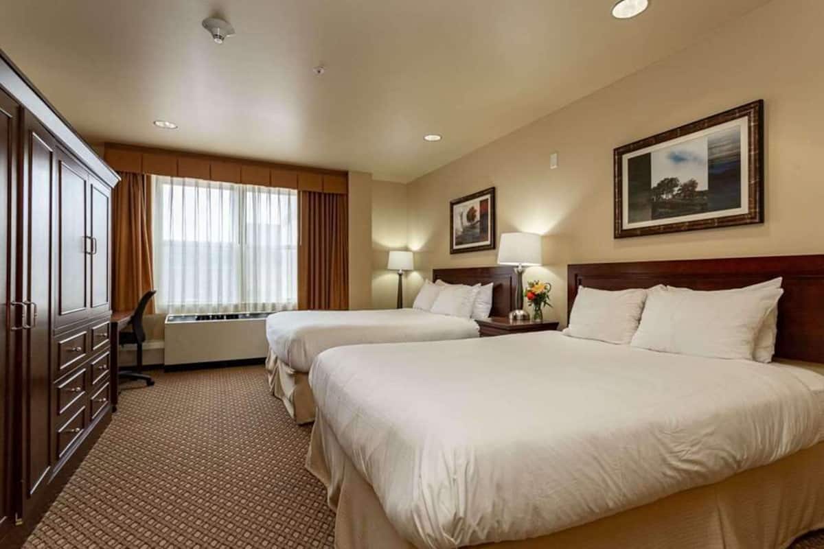 A hotel room with two beds, a desk, and a chair. There are two windows with curtains, a radiator, and a framed picture on the wall.