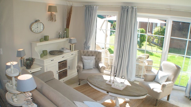 A cozy living room with a sofa, armchairs, a coffee table, and a dining table. There is a window with curtains, a clock on the wall, and a lamp on the side table.