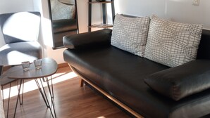 A modern living room with a black leather sofa, a round wooden coffee table, and a small side table. There is a black metal ladder with shelves against the wall, and a large mirror above it. - 3-room guest apartment close to the center, suitable for 1 - 4 persons (Leipzig)