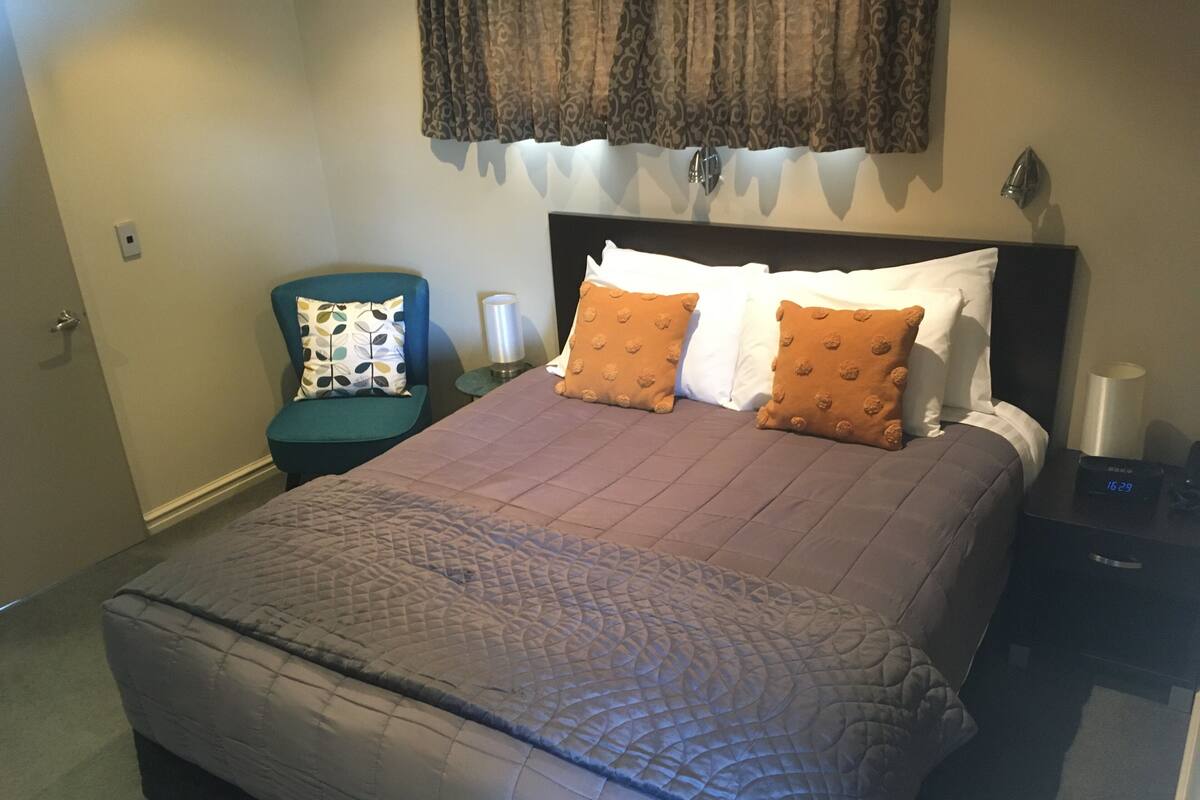Una cama bien hecha con una colcha estampada, dos almohadas con diseño texturizado, una mesita de noche con una lámpara y un reloj de pared.