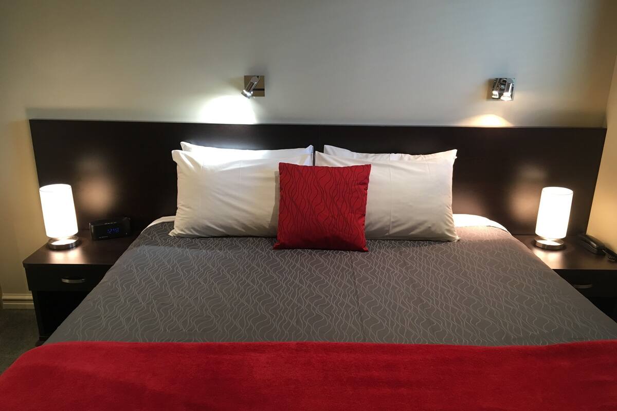 Una cama bien tendida con una colcha estampada, dos lámparas de noche, un reloj digital y un cojín decorativo rojo.