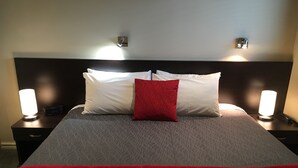 A neatly made bed with a red pillow, two bedside lamps, and a digital clock on a bedside table. - Kauri Motel (Christchurch)