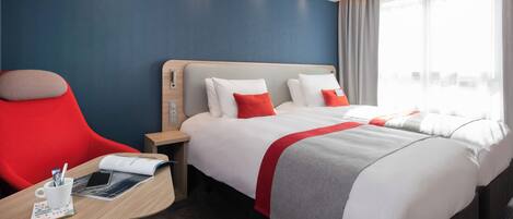 Una habitación de hotel con una cama, un sillón rojo, una mesita de noche con un libro y una taza, y una ventana con cortinas.