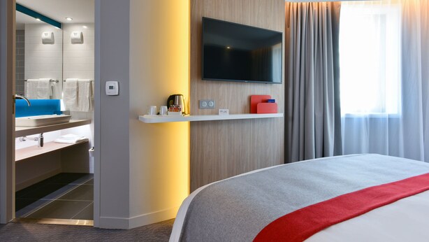 Une chambre d’hôtel avec un grand lit, un téléviseur fixé au mur, une salle de bain avec un miroir et une fenêtre avec des rideaux.