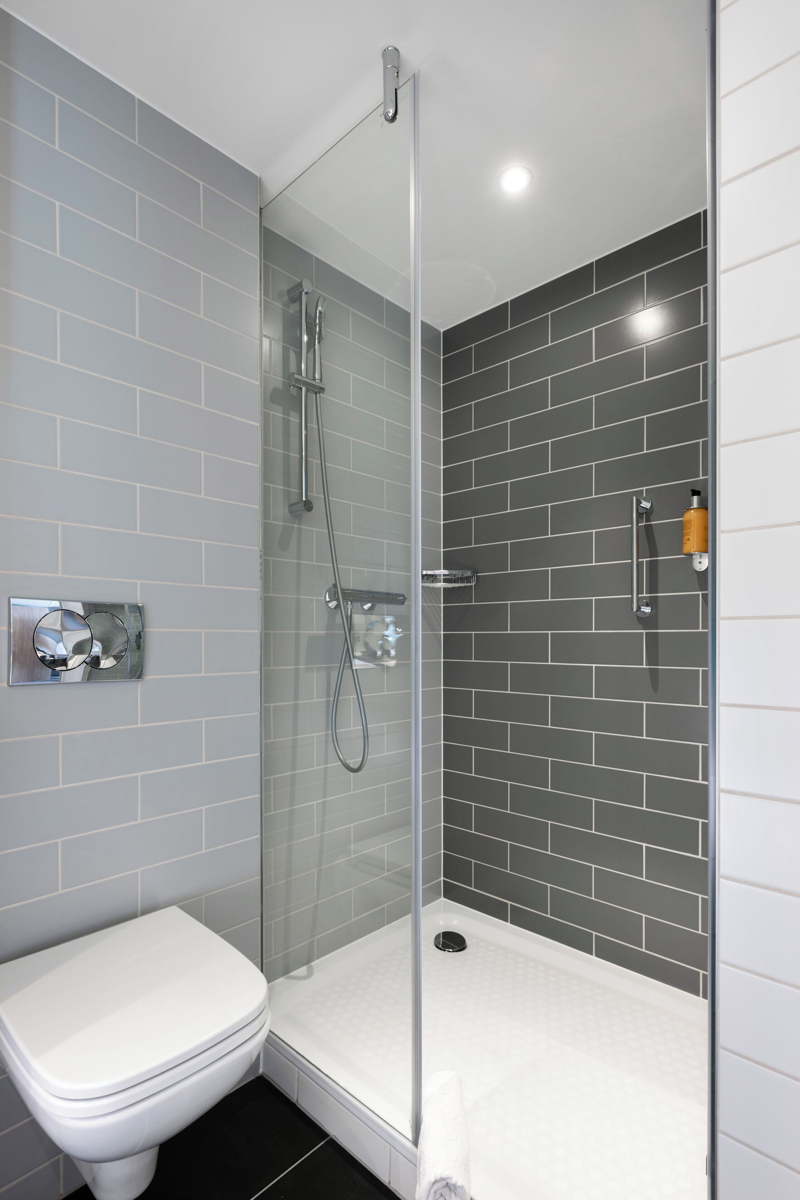 Un baño moderno con ducha envuelta en vidrio, inodoro blanco y paredes con azulejos grises.