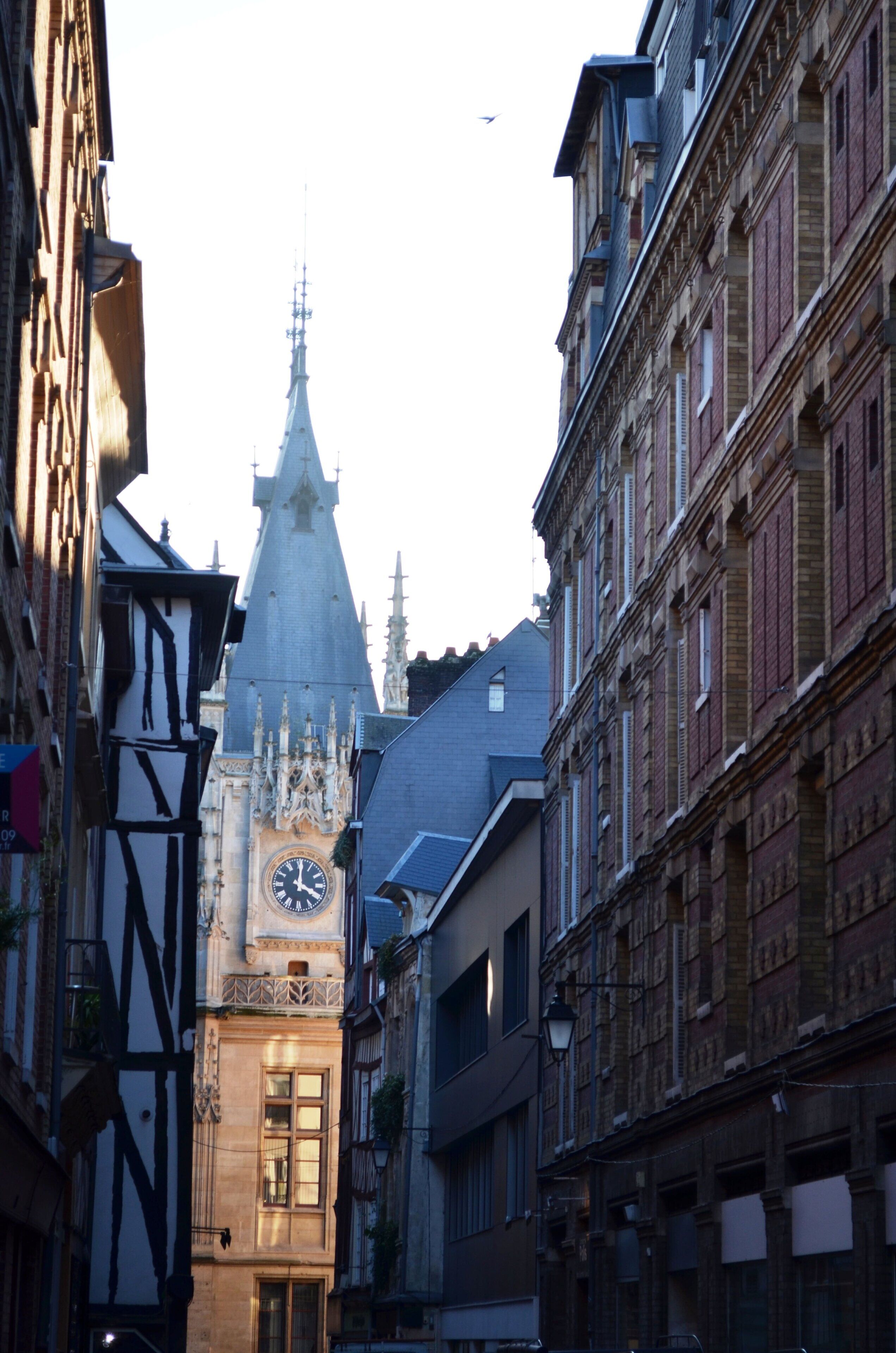 Una calle estrecha con edificios históricos, una imponente torre del reloj y la aguja de una iglesia.