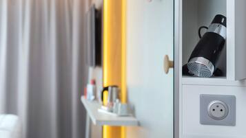 A hotel room with a bed, a bedside table with toiletries, a mirror, and a wardrobe with a hanging iron and a vacuum cleaner.
