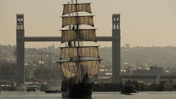 Un barco de velas alto con varias velas en un puerto, con un puente y edificios al fondo.