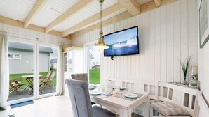 A cozy room with a wooden ceiling, a television mounted on the wall, a dining table set with plates and cups, a cushioned bench, and a view of a house and lawn outside.