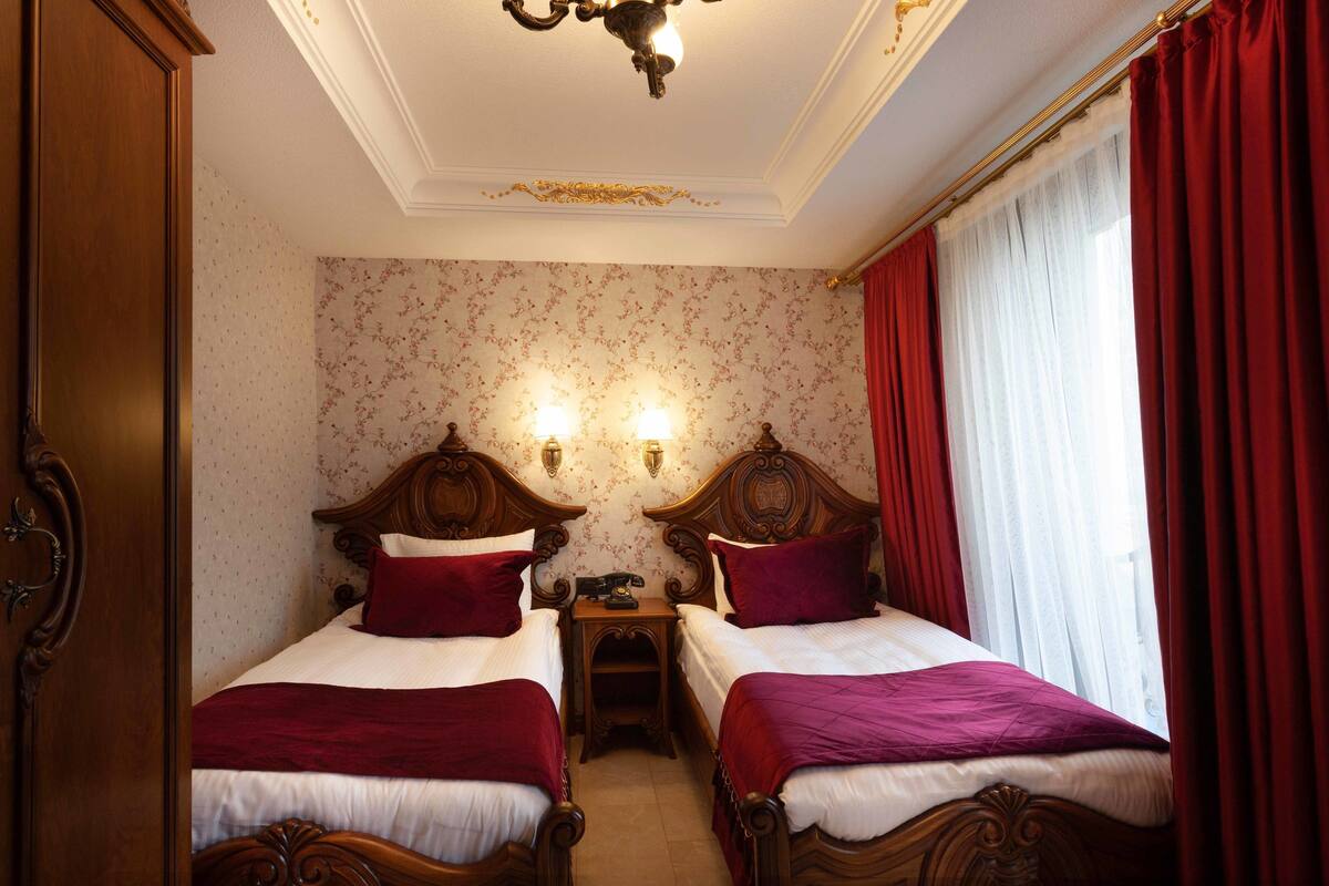 A hotel room with two wooden beds, red curtains, floral wallpaper, and a chandelier.