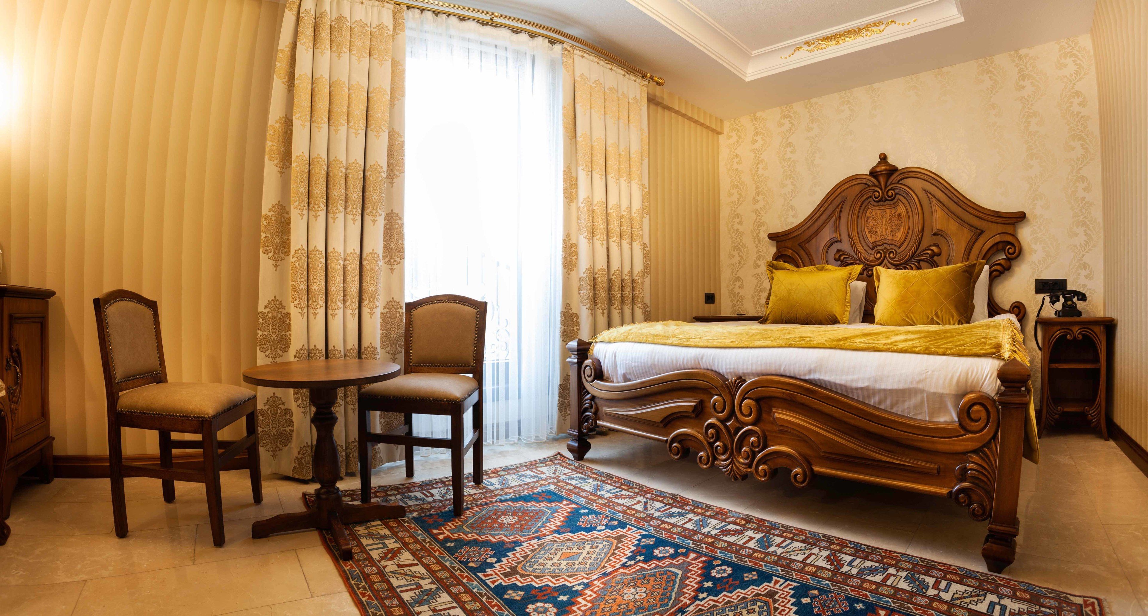 Un dormitorio con una cama grande, una silla de madera, una mesita pequeña y una alfombra estampada.
