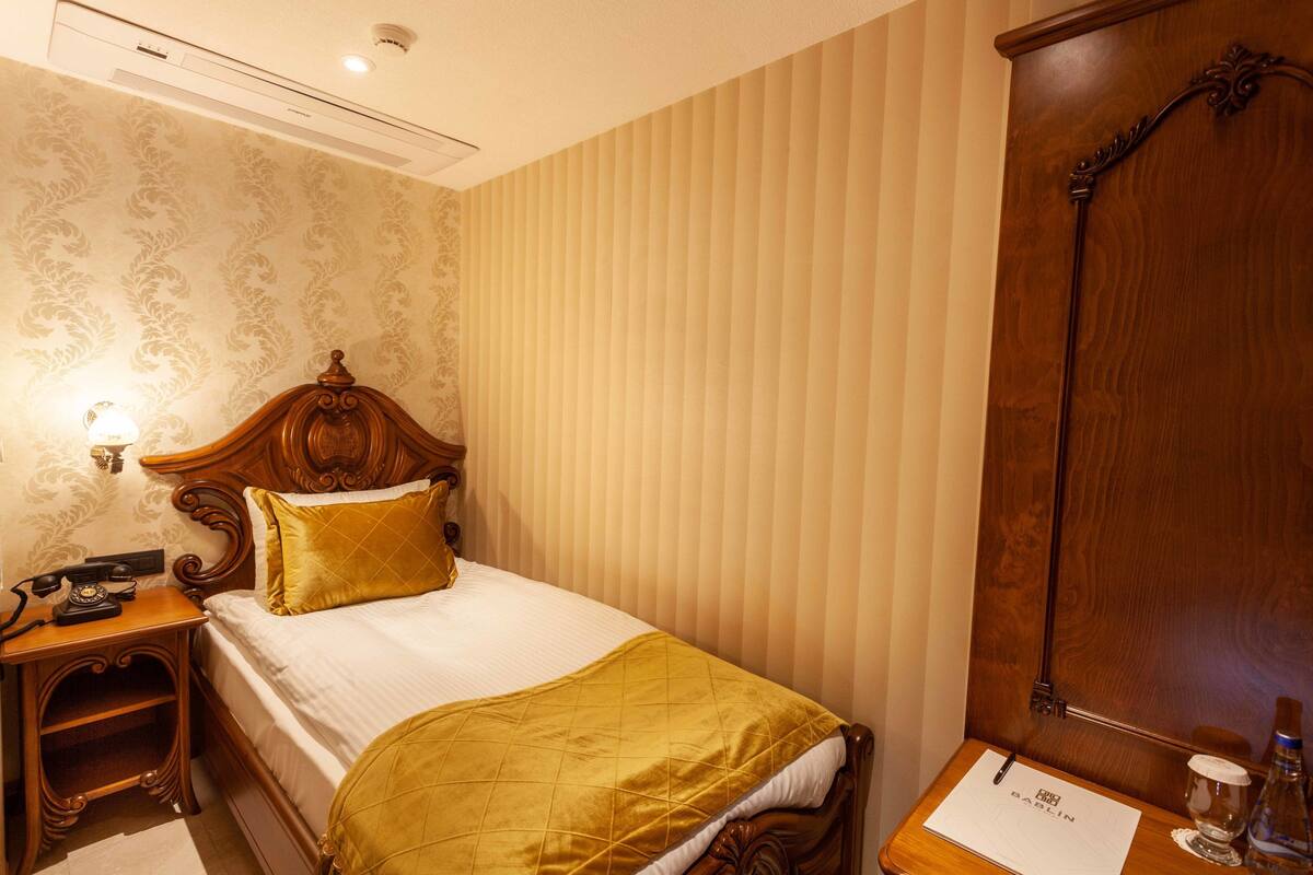 A hotel room with a wooden bed, a nightstand with a telephone, a bedside table with a lamp, and a wooden wardrobe.