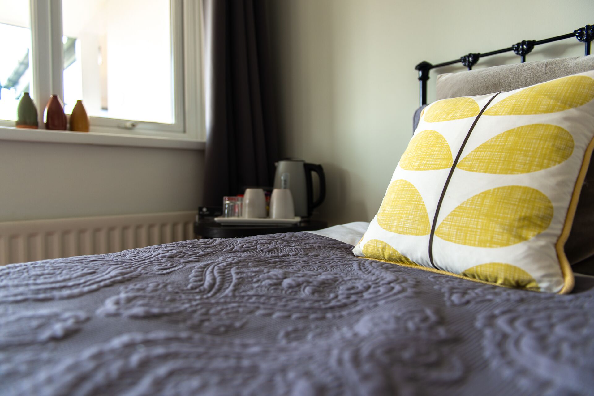 Una cama bien hecha con una colcha estampada, un almohadón con diseño amarillo y blanco, y una ventana con cortinas.