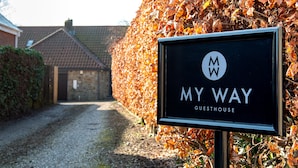 A sign for 'MY WAY GUESTHOUSE' with a logo, set against a backdrop of autumn leaves and a gravel path leading to a building. - My Way Guest House (Durham)