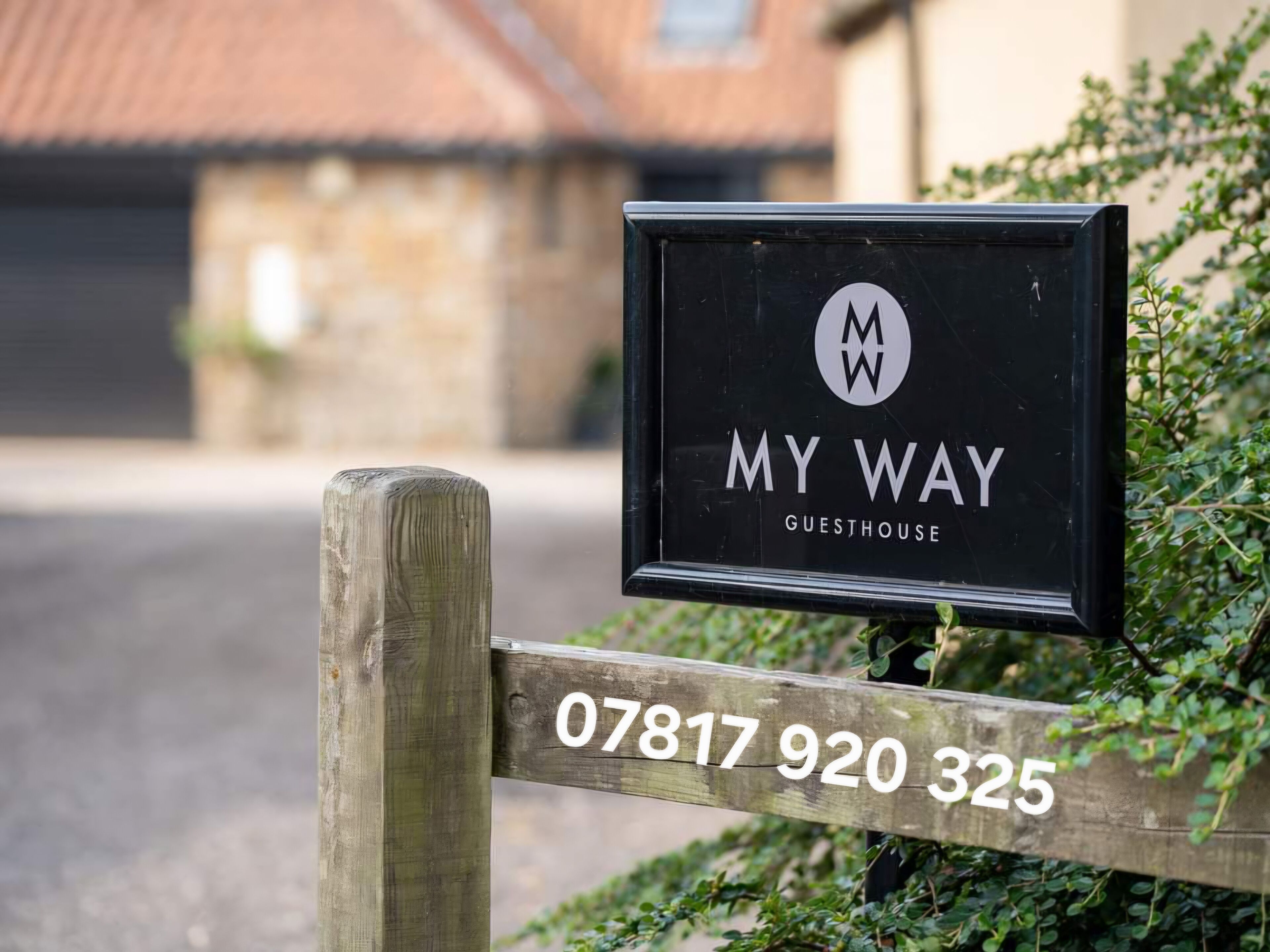 A black sign with 'MY WAY GUESTHOUSE' and a phone number '07817 920 325' on a wooden post.