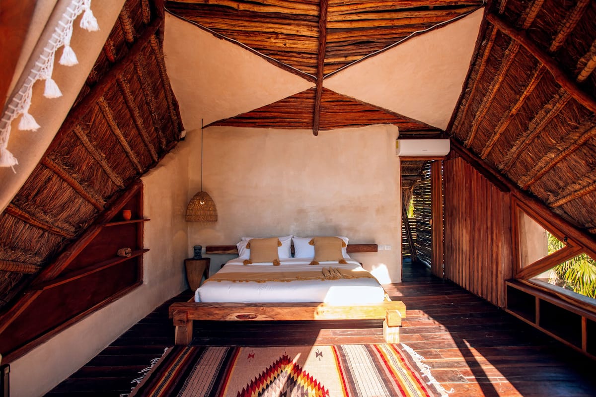 A spacious room with a large bed, wooden beams, and a view of greenery outside.