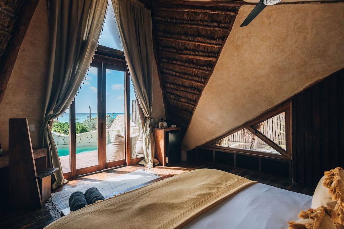 A bedroom with a large window, wooden beams, a bed, and a view of the sea.