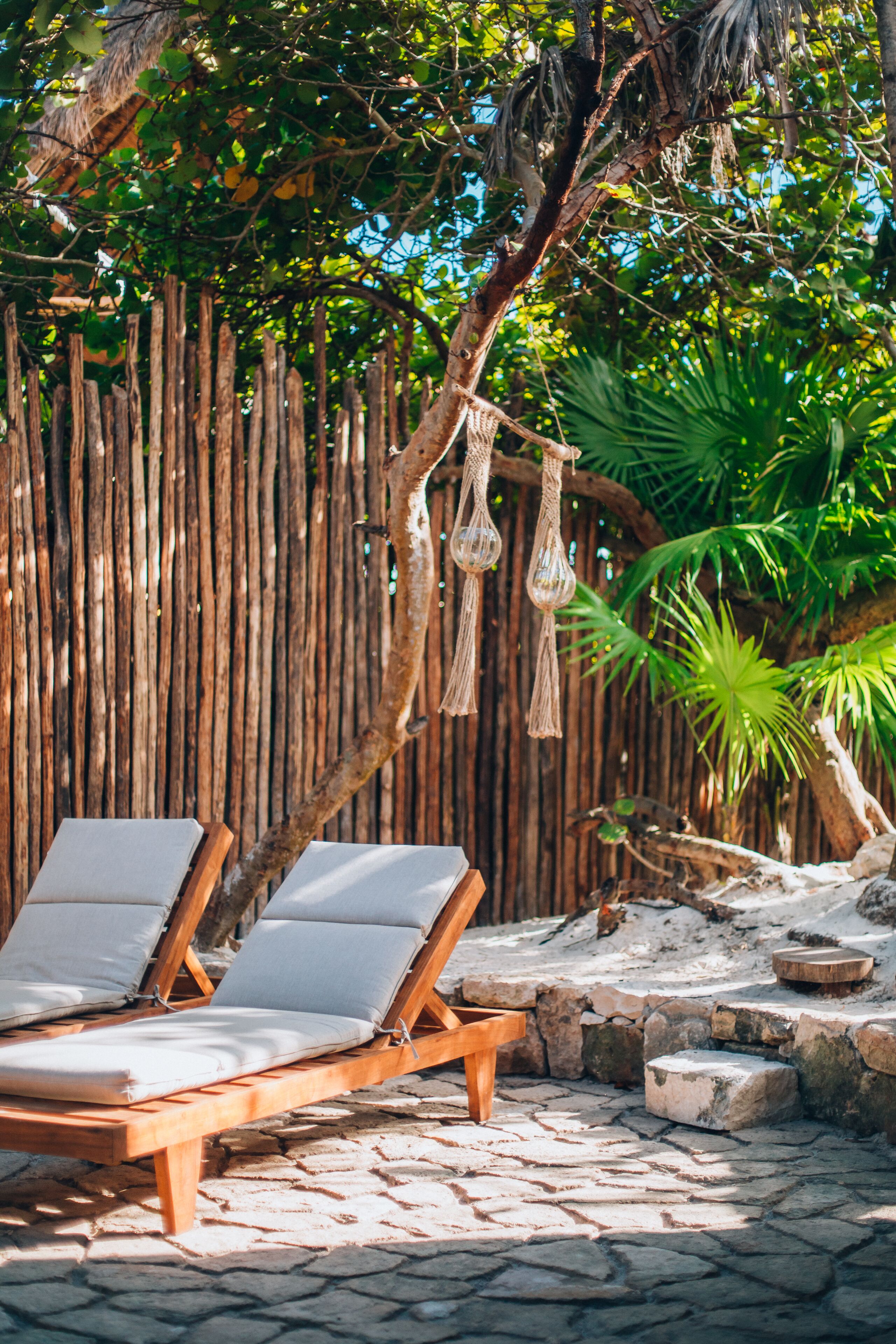 A wooden lounge chair with cushions, a stone pathway, a wooden fence, and hanging macramé decorations.