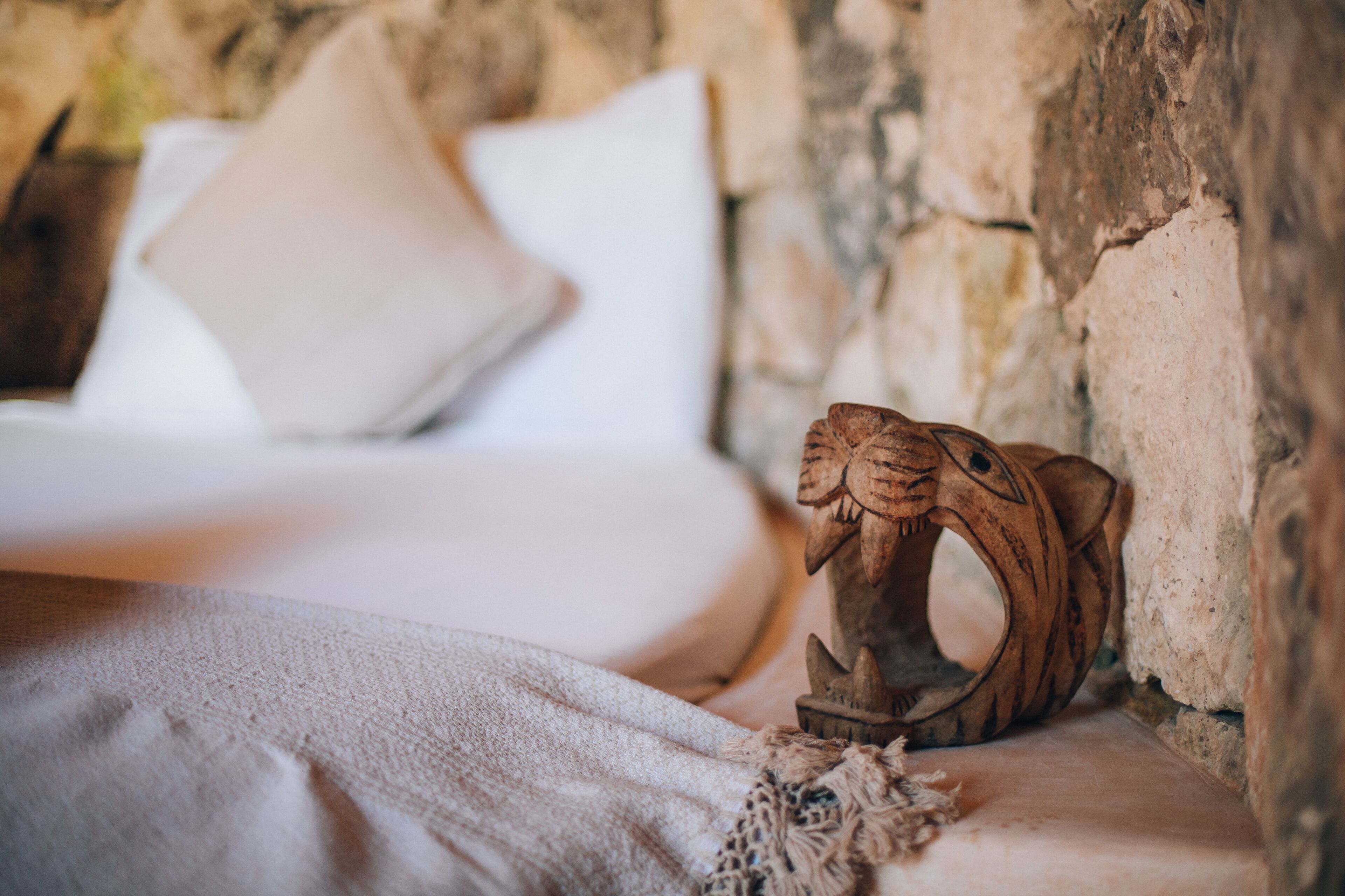 A bed with white linens and a beige pillow, a wooden sculpture on the side, and a stone wall in the background.