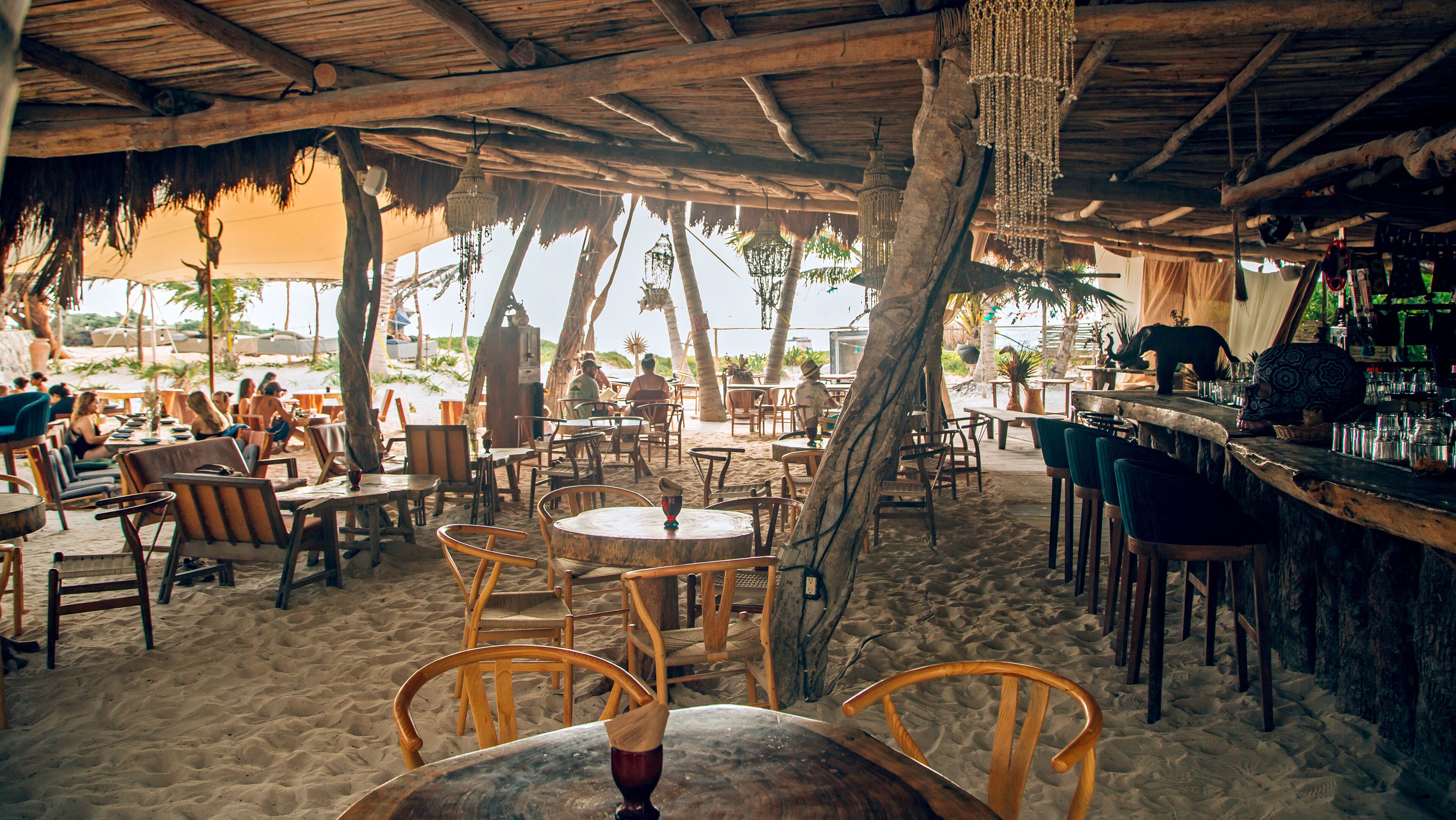 An outdoor restaurant with wooden tables and chairs, a bar area, and a view of the beach.