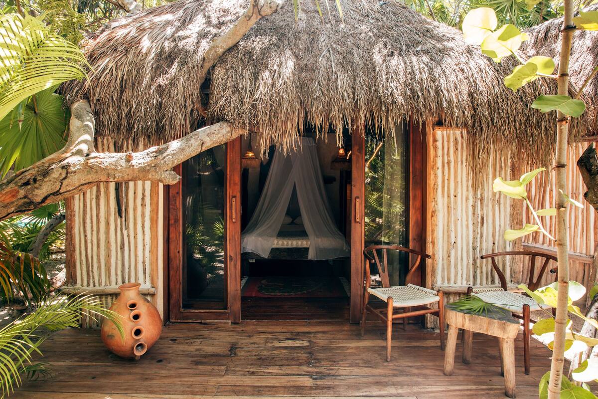 A thatched-roof hut with a bed, wooden chairs, and a clay pot.