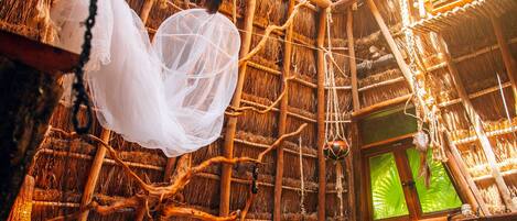 A thatched-roof room with a bed, hanging net, and a window with a view.