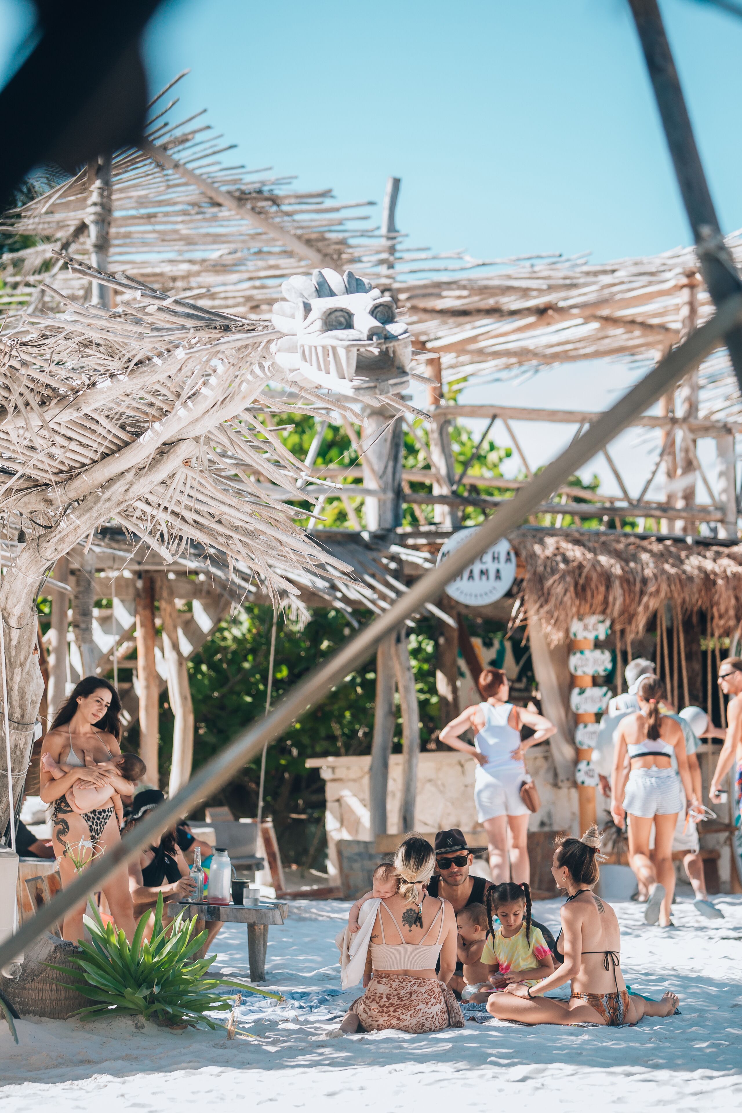 A beach scene with people relaxing under a thatched structure, a sign reading "Mochila Mama", and a table with drinks.