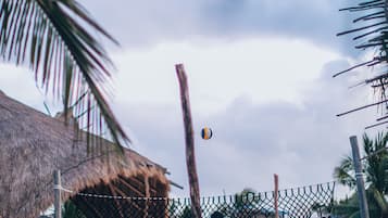 Un partido de voleibol de playa con jugadores en shorts, una estructura de techo de paja y palmeras al fondo.