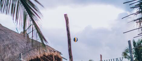 Un partido de voleibol de playa con jugadores en shorts, una estructura de techo de paja y palmeras al fondo.