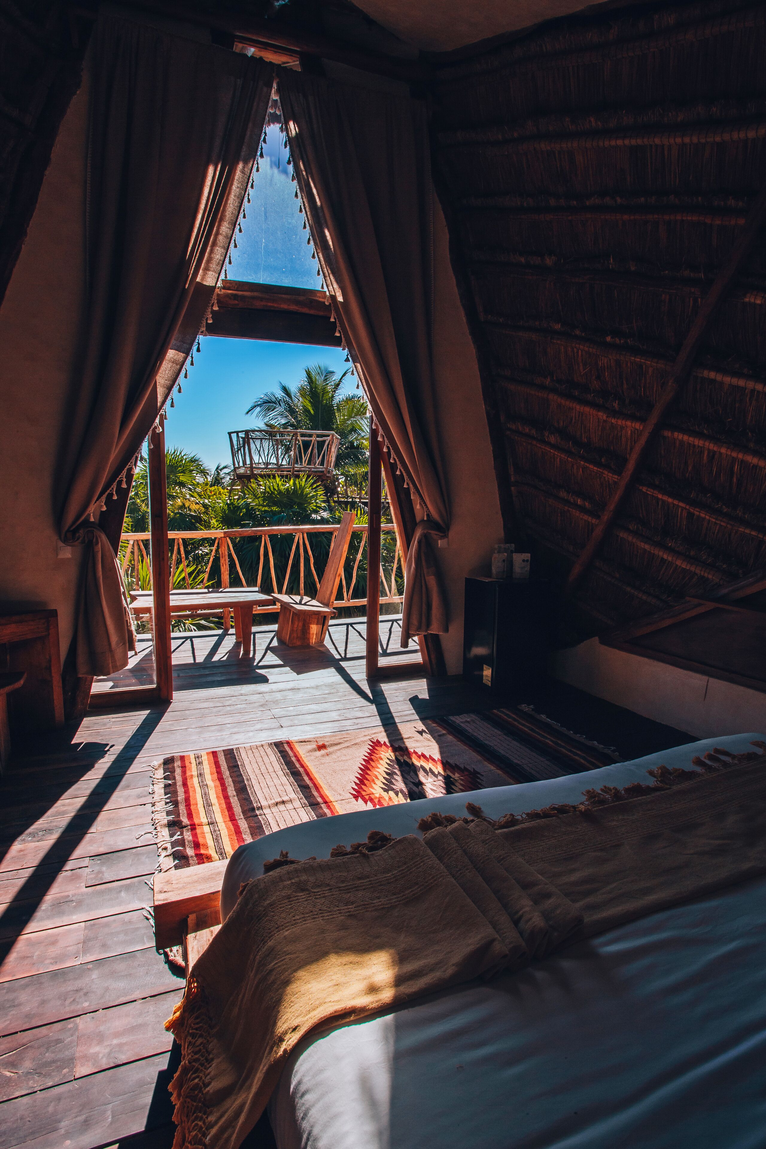 A room with a bed, wooden floor, curtains, a window, and a view of palm trees.