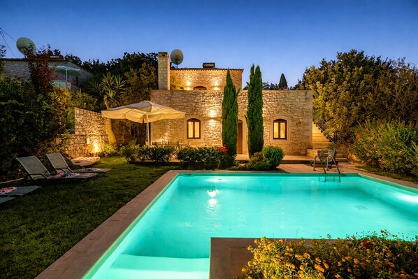 A stone villa with a swimming pool, outdoor seating, and a well-lit garden at dusk.