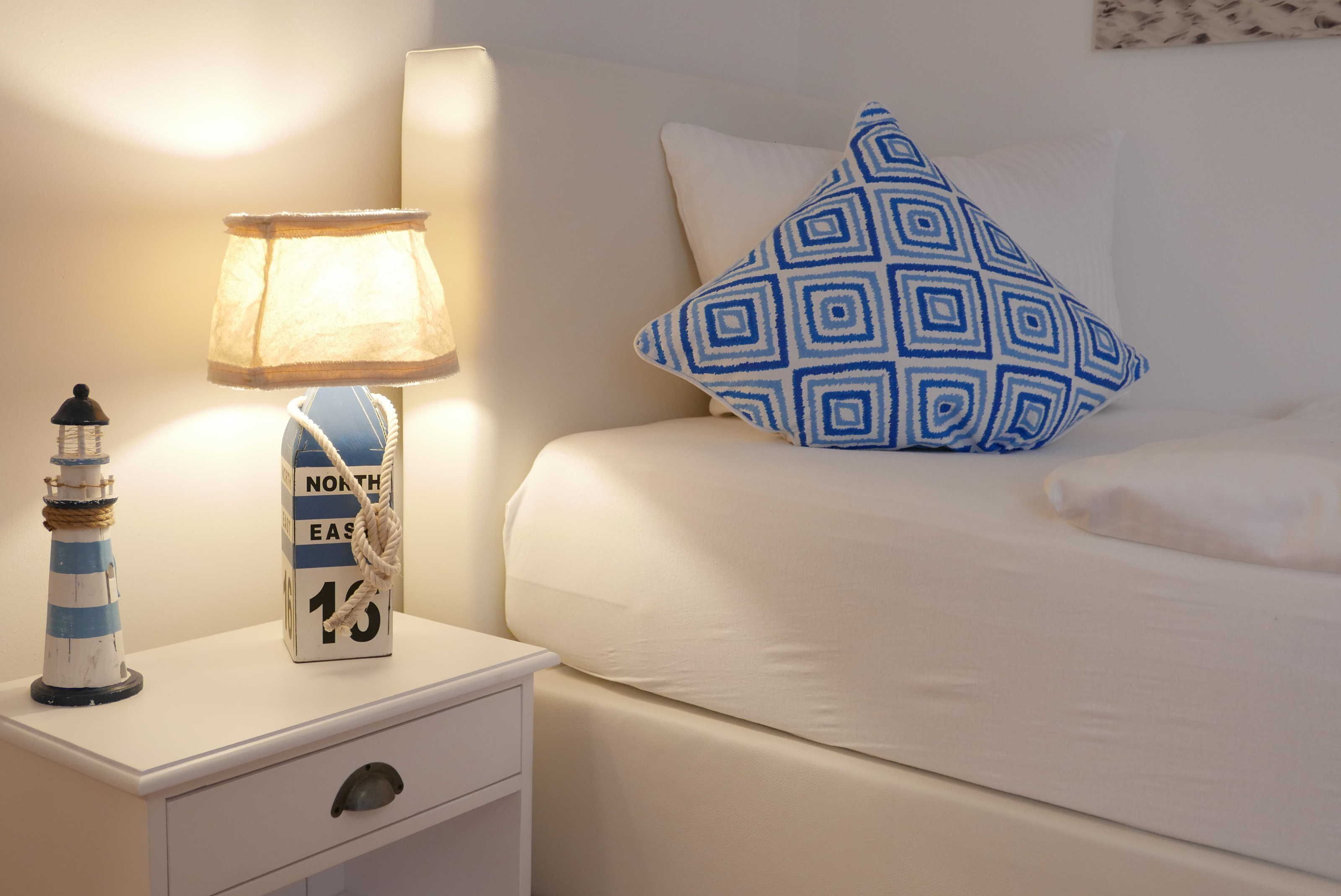 Un lit bien fait, agrémenté d’un coussin à motifs bleus, une table de chevet blanche avec une lampe et une petite décoration représentant un phare, et un luminaire fixé au mur.