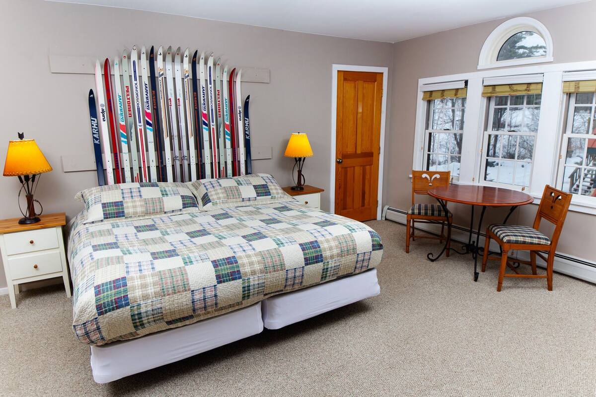 A bedroom with a bed, two bedside tables, a lamp, a wooden chair, a dining table, and a window with a view of trees.