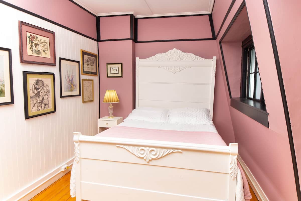 A pink-themed bedroom with a white bed, framed pictures on the wall, and a window with a view.