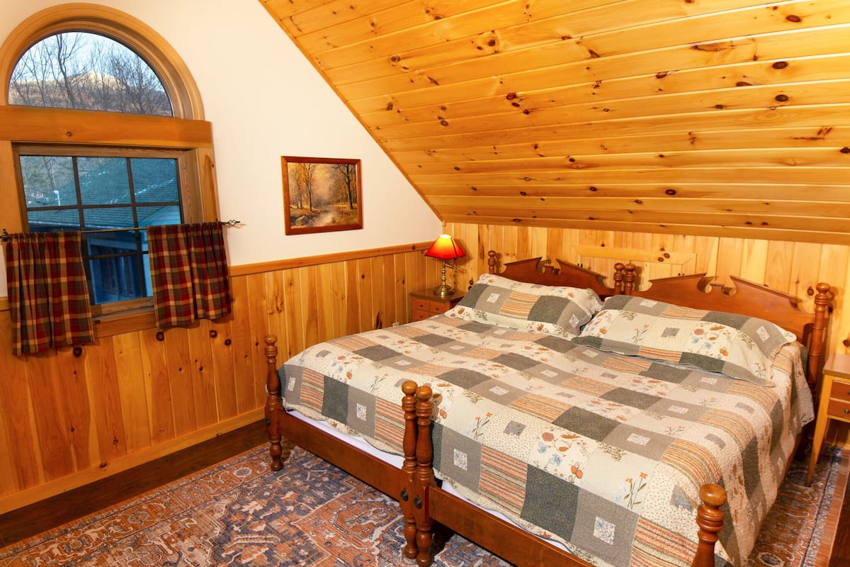 A bedroom with a wooden ceiling, a large bed with a quilt, a window with plaid curtains, and a wooden floor.