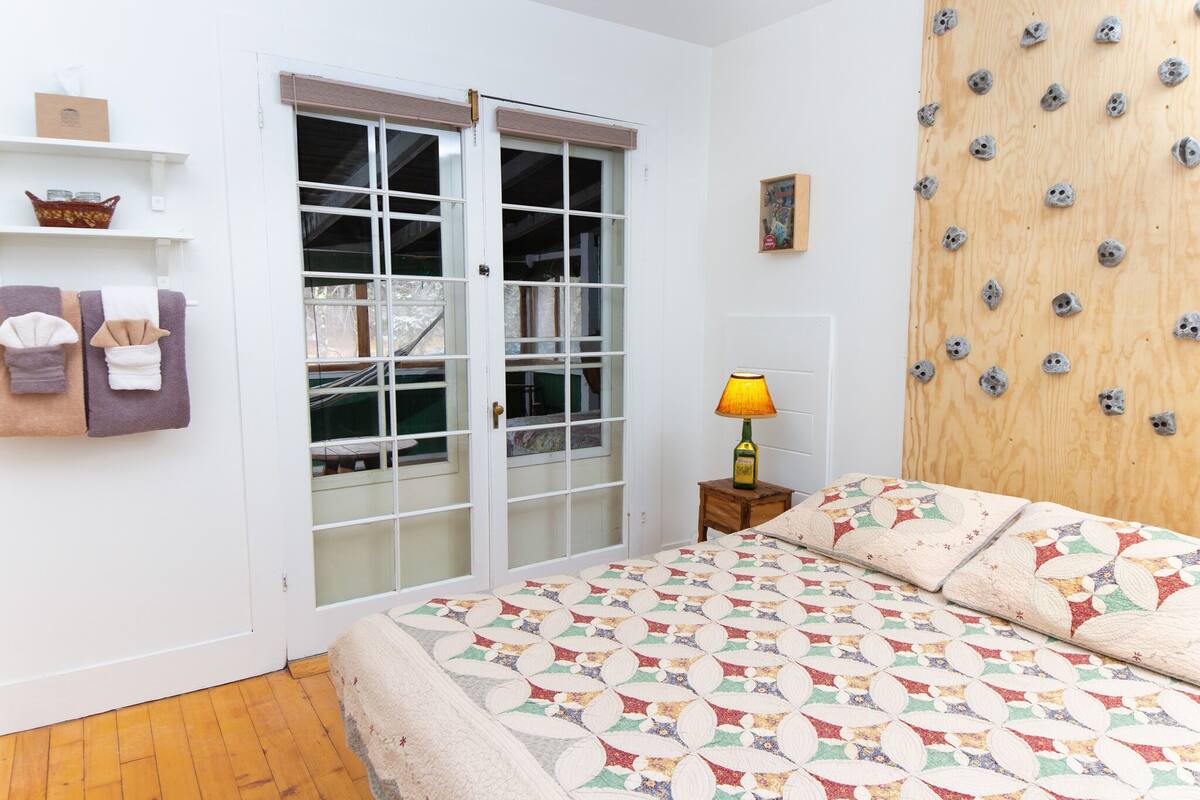 A bedroom with a bed, a wooden wall with pegs, a window with a view, and a door.