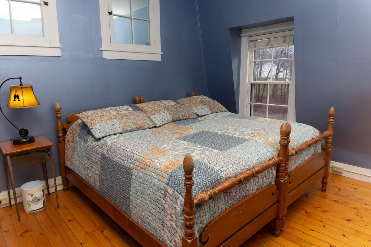 A bedroom with a wooden bed, a nightstand with a lamp, and a window with a view of trees.
